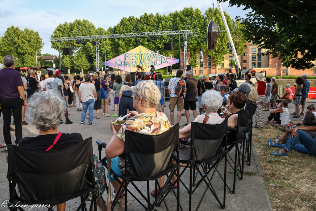 Photo de personnes assises dans des chaises pliables avec une assise haute, des accoudoirs et des dossiers qui regardent et écoutent un concert du festival Convivencia.
